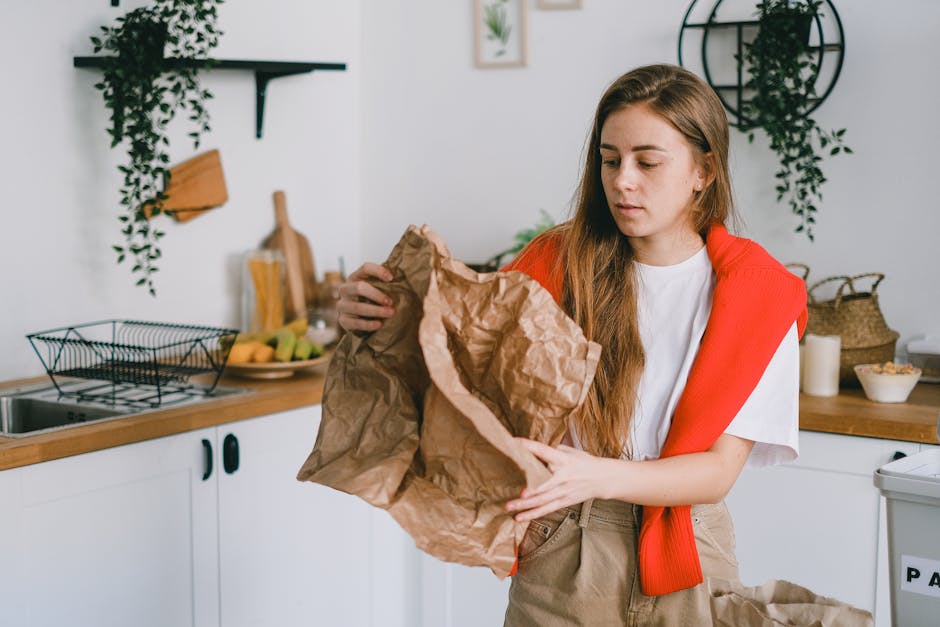 A young woman with long brown hair, wearing a white t-shirt and a red sweater draped over her shoulders, stands in a modern kitchen with white cabinets and wooden countertops. She is holding a large crumpled brown paper bag with both hands, examining it closely. Behind her, there are decorative green plants hanging on the wall, a few framed pictures, and a wire dish rack on the countertop with a wooden cutting board and a bowl of fruit. To her right, there is a white rubbish bin with a partially visible black label. The environment looks clean and organized, with natural lighting illuminating the scene, which relates to waste management and the preparation of household rubbish for disposal or collection, hinting at a focus on domestic rubbish removal services.