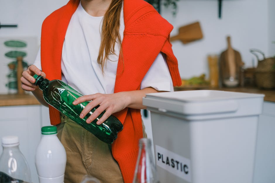 The image features a person in a kitchen or indoor environment, with only the upper body partially visible. They are wearing a white t-shirt and have an orange knitted sweater draped over their shoulders. The individual is holding a green plastic bottle with a black cap, examining or preparing to recycle it. In front of them, there is a white recycling bin labeled 'PLASTIC,' positioned on a countertop or table, which appears to be in a domestic or small-scale collection setting. The background includes various kitchen items, such as bottles, containers, and a wooden shelf with utensils, suggesting a space dedicated to household activities related to waste sorting or recycling. The scene is illuminated with natural or soft lighting, emphasizing a neutral, practical atmosphere suitable for discussing waste management or private rubbish collection services like those offered by Junk Removals Services, which specialize in alternative waste handling options outside of local authority collections.