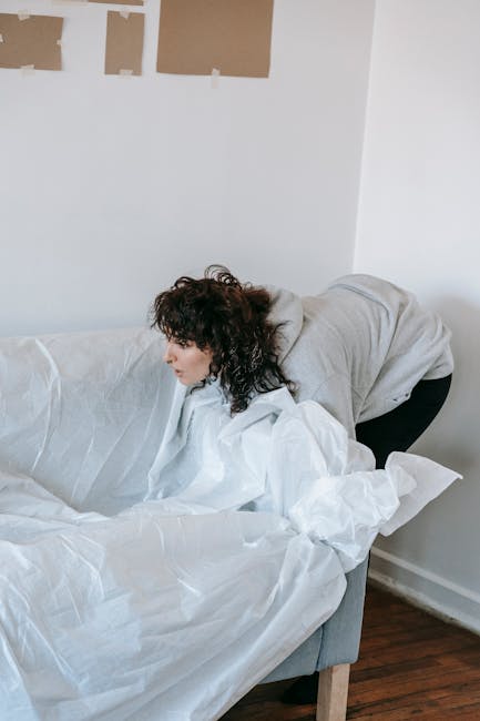 A woman with shoulder-length curly dark hair, wearing a light grey hoodie and black pants, is seen leaning over the arm of a plain light grey upholstered sofa, engaged in cleaning or rearranging. The 