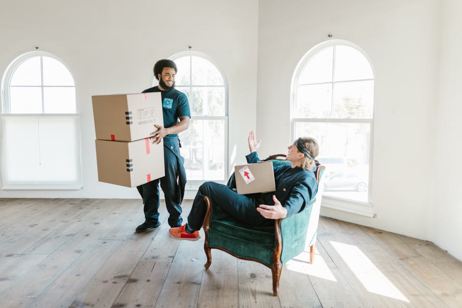 A woman with shoulder-length curly dark hair, wearing a light grey hoodie and black pants, is seen leaning over the arm of a plain light grey upholstered sofa, engaged in cleaning or rearranging. The 