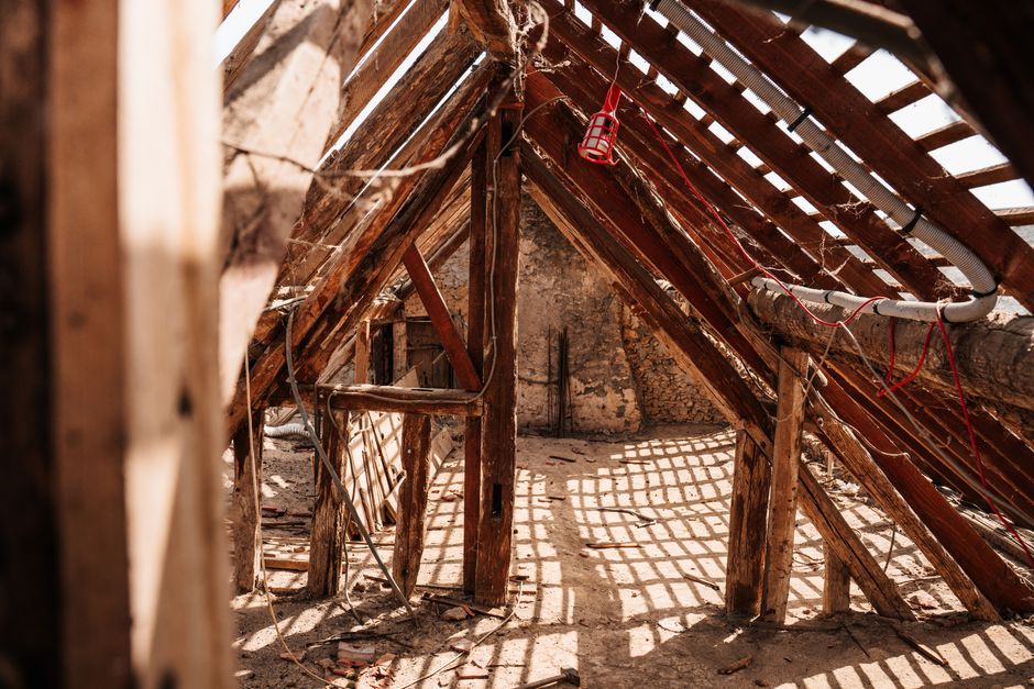 Photograph of the interior of an old, partially collapsed brick barn or loft space with a ceiling open to the sky. The brick walls are weathered and reddish-brown, and some hay or straw remnants are s